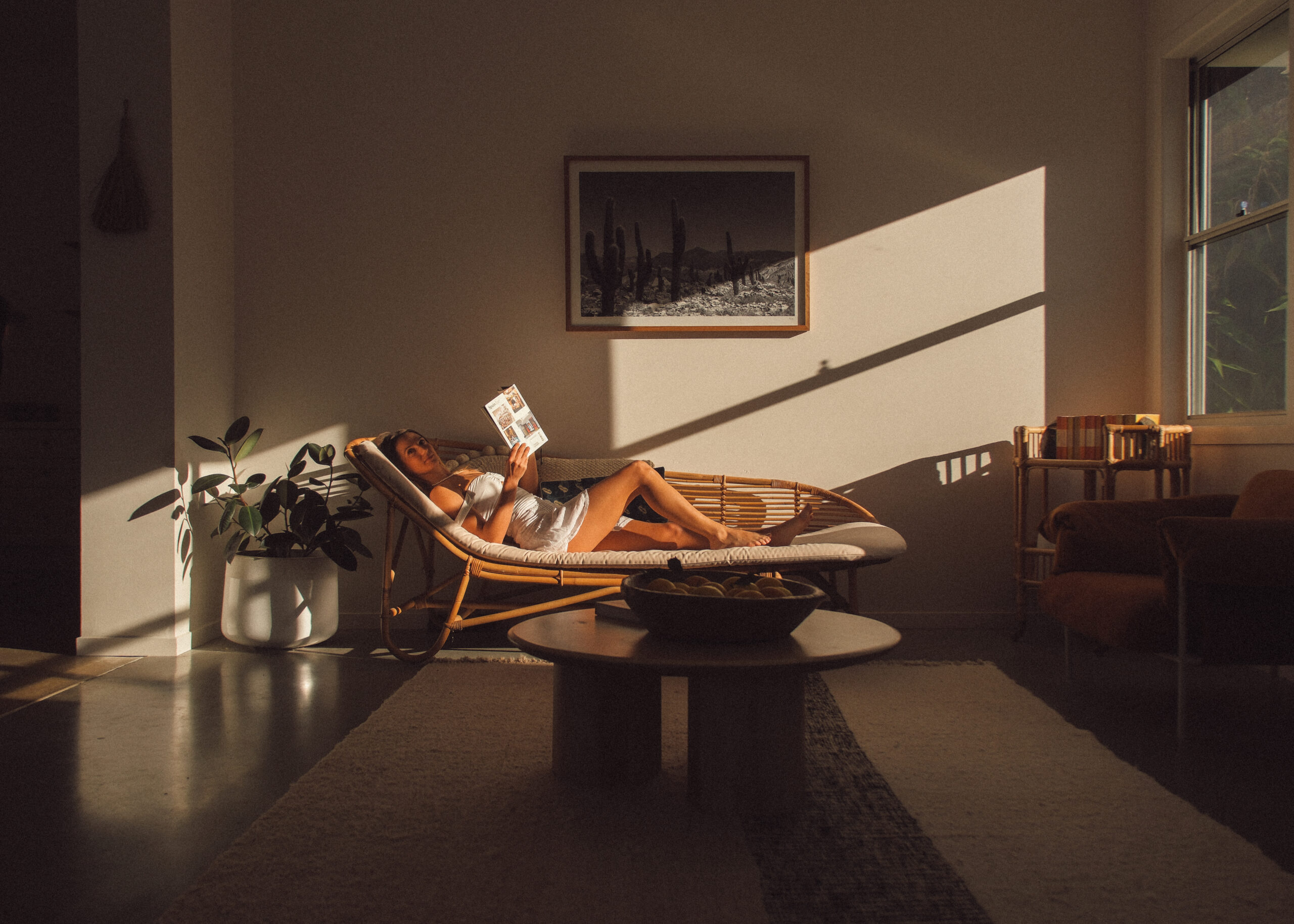 Woman reclining inside Bailen Byron’s lounge, sunlight casting across the wall, framed by potted plants and rattan.