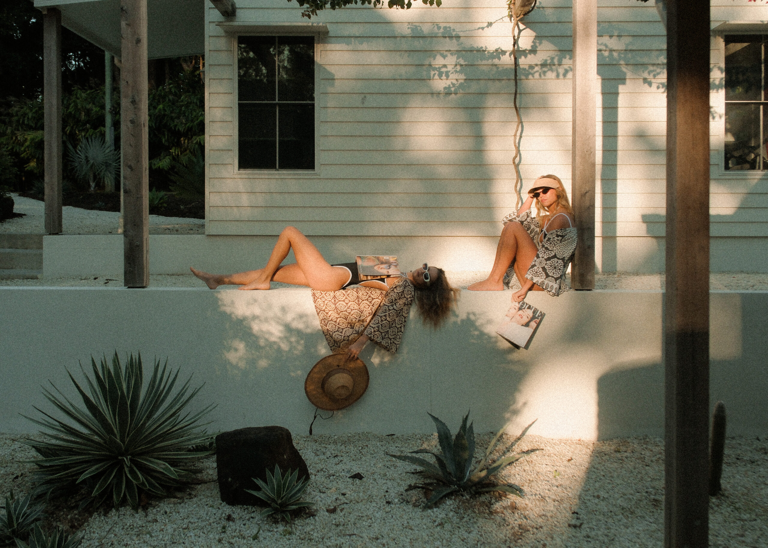 Two women lounging on a white rendered terrace, reading magazines in the afternoon sun at a villa in Ewingsdale.
